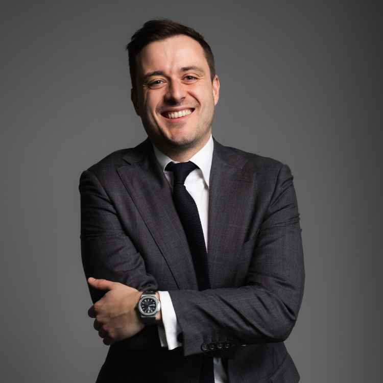 Tom McKinnon headshot on a grey background - he is wearing a grey suit and a white shirt smiling.
