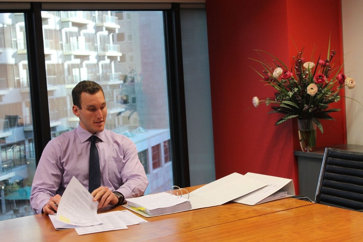 Eleven years on: how National Compensation Lawyers began - Managing Principal Michael Tanner at his first desk, in a light purple shirt and tie, sitting at a wooden desk covered in legal documents and folders during the early days of National Compensation Lawyers.